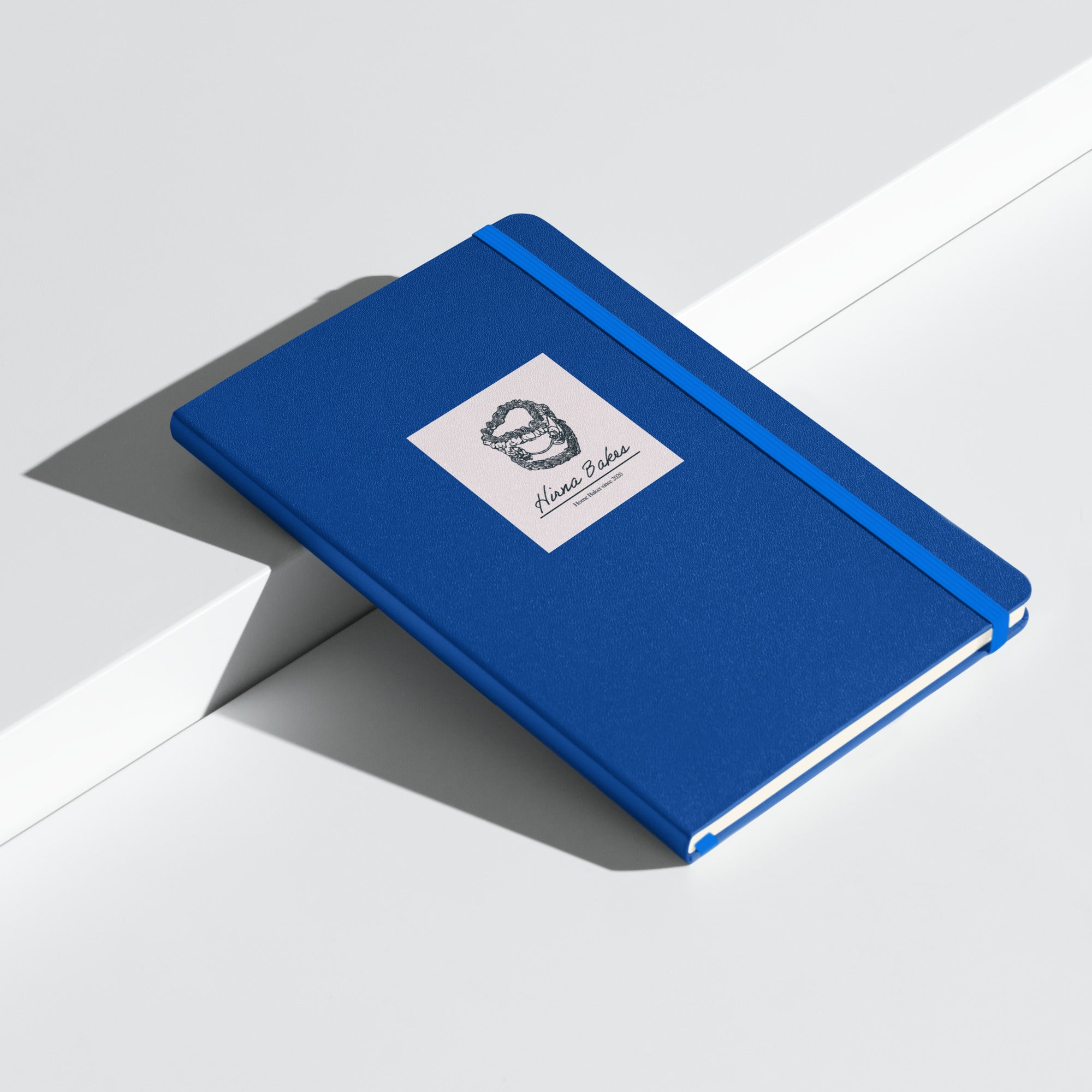 Blue hardcover Hirna Bakes notebook laying flat at an angle on a white surface